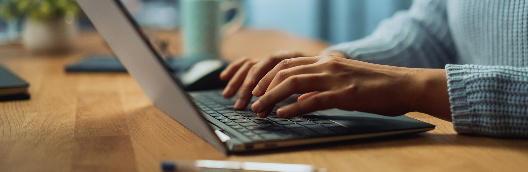 Close-up of a person typing on a laptop keyboard, seated at a wooden desk. The person is wearing a light blue sweater, and there is a coffee mug and notebook on the desk in the background