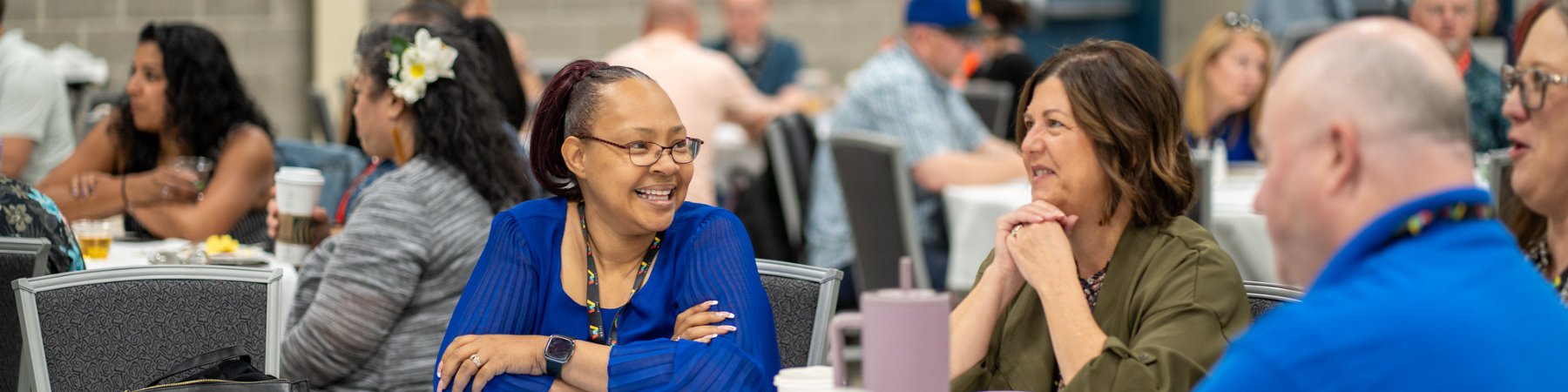 Groups of people at tables featuring a black woman in the center with a blue shirt smiling at others at the table.