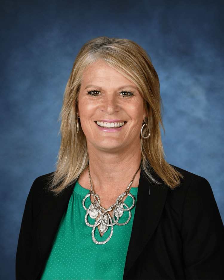 A smiling woman with shoulder-length blonde hair wearing a green blouse, a black blazer, and a large silver necklace, set against a blue background.